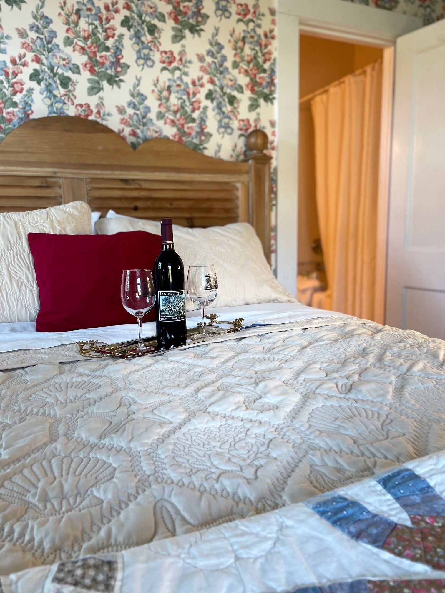 Una cama con cabecera estampada de flores, almohada roja y una botella de vino con una copa.