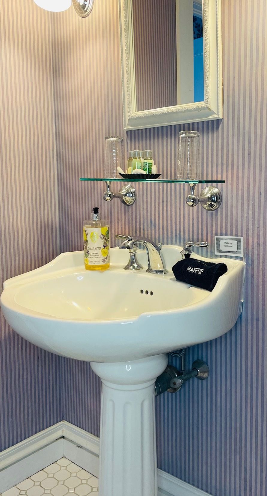 A bathroom with a white pedestal sink, a glass shelf above the sink, and a mirror with a decorative frame.