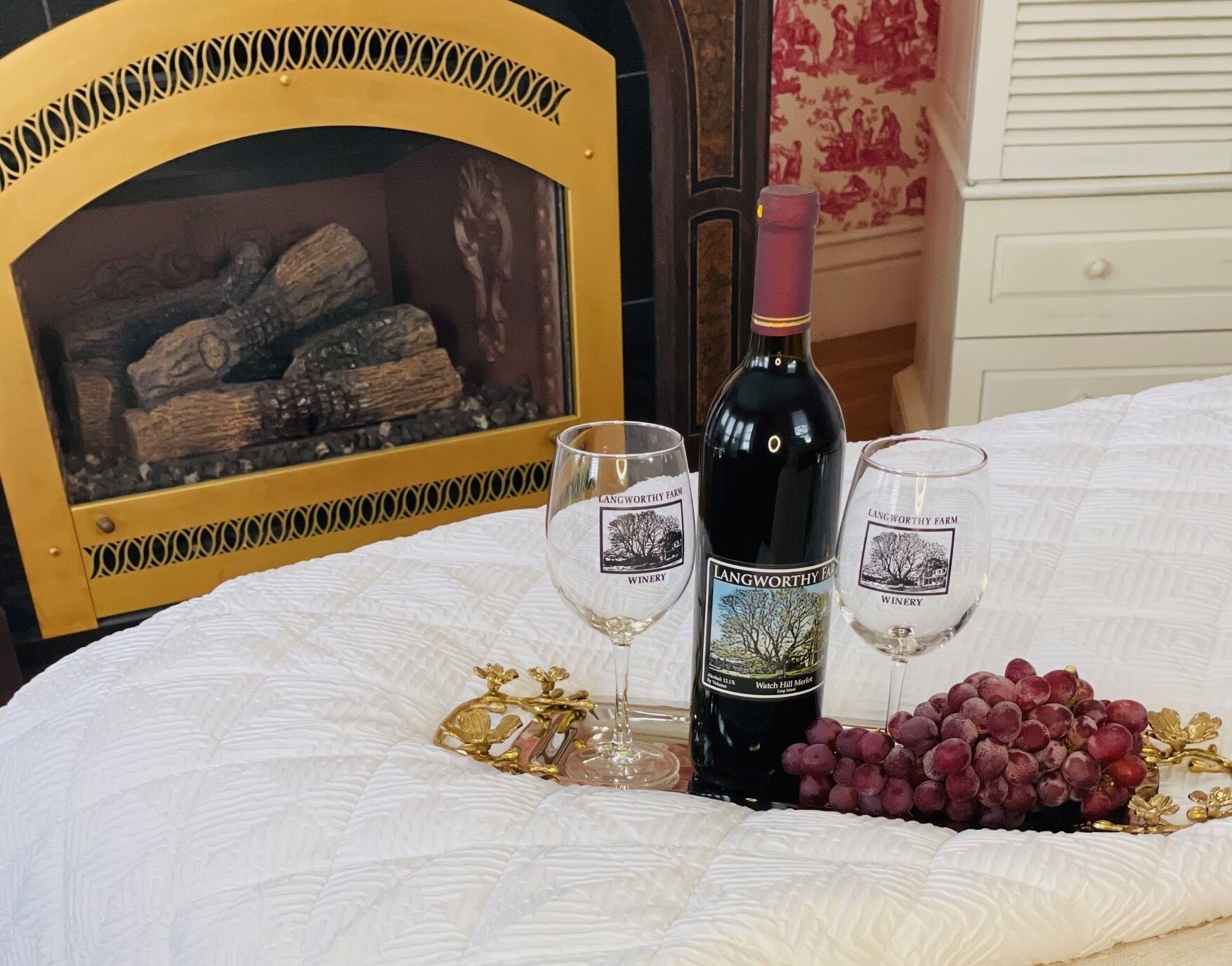 A bed with a white quilt, a bottle of wine, two wine glasses, and a bunch of grapes on a table.
