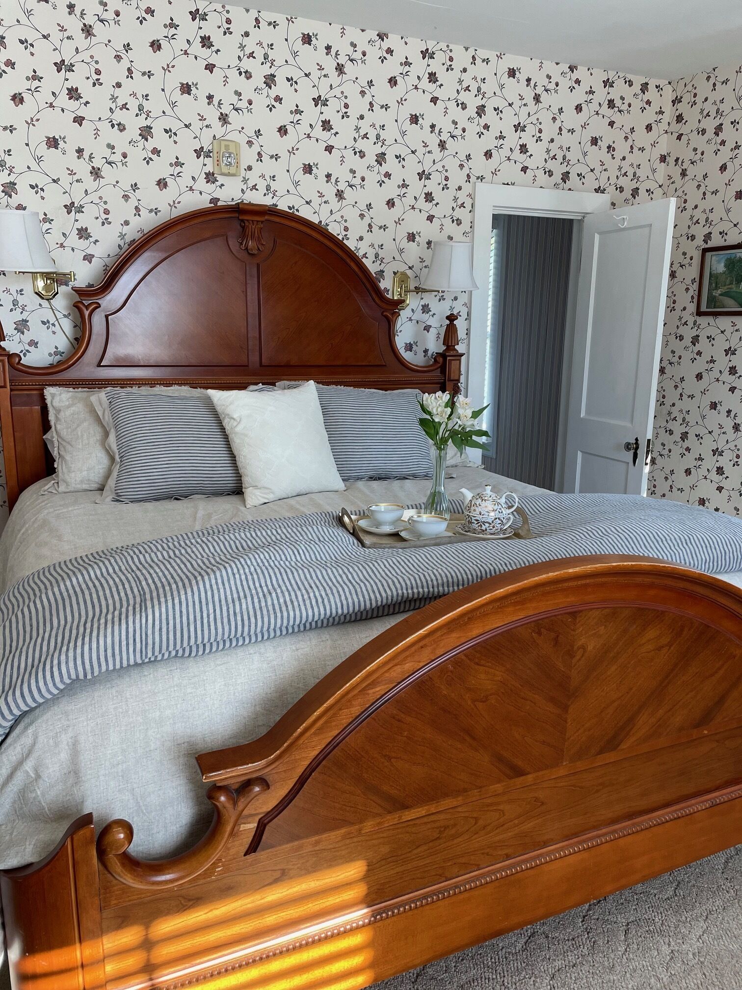 Une chambre à coucher avec un lit en bois, un papier peint à motifs floraux et une porte sur le côté droit.