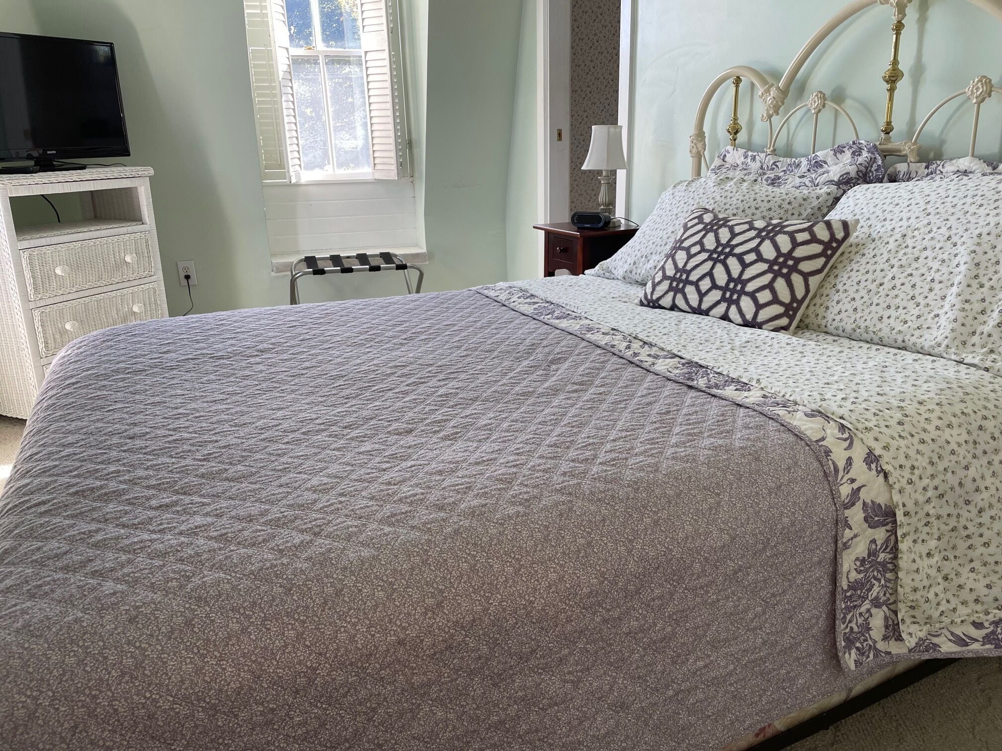 A neatly made bed with a patterned bedspread and pillows, a bedside table with a lamp, and a television on a stand.
