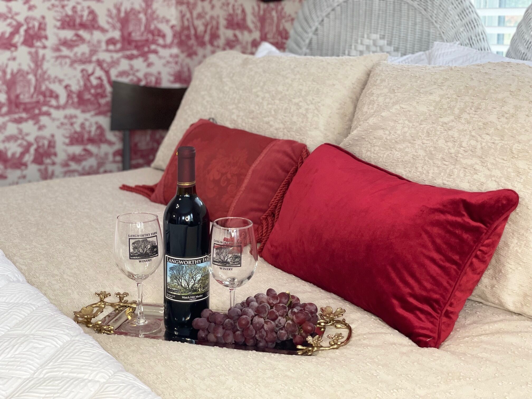 A bed with a wine bottle, two wine glasses, and a bunch of grapes on a tray.