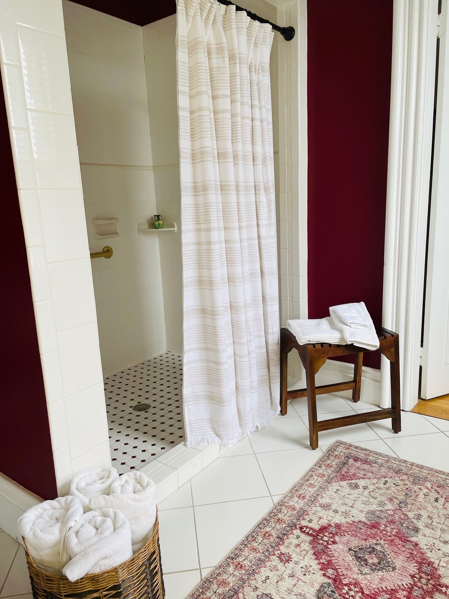A bathroom with a shower, a wooden stool, a patterned rug, and a basket of towels.
