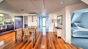 A modern hotel room with a wooden floor, a dining area with wicker chairs, a bed with a blue and white striped duvet, and a kitchenette with a refrigerator and cabinets.