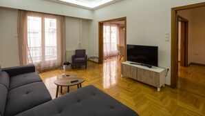 Een moderne woonkamer met een grijze bank, een tv op een houten dressoir, en een zichtbare eethoek door een deuropening.