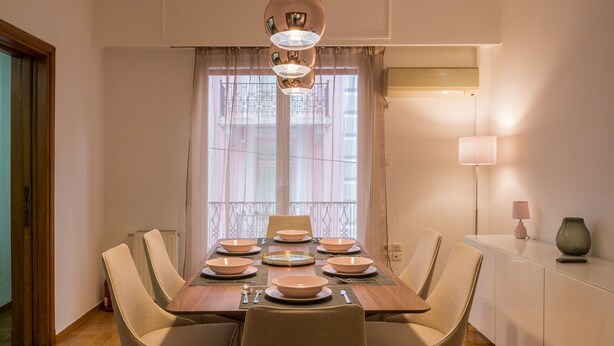 Een eetgedeelte met een houten tafel voor zes personen, zes stoelen, een kroonluchter en een raam met gordijnen.
