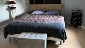 A bedroom with a bed, bench, bedside table, lamp, and a small black storage unit. - Furnished with charm in the Drôme Provençale (La Bégude-de-Mazenc)