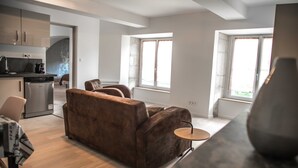 A modern living room with a brown sofa, a small round table, and a kitchen area with cabinets and appliances. - Furnished with charm in the Drôme Provençale (La Bégude-de-Mazenc)