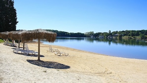A beach with thatched umbrellas and lounge chairs, a calm lake, and houses in the background.
