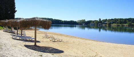 Una playa con sombrillas de paja y sillas de descanso, un lago tranquilo, y casas al fondo.