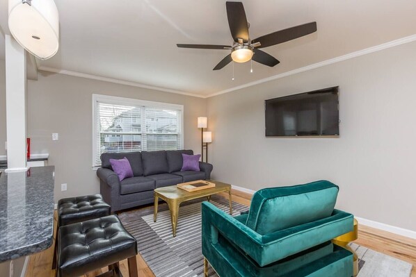 A living room with a sofa, a teal armchair, a coffee table, a ceiling fan, and a TV mounted on the wall. - Trendy 1 BR w/KING Bed in Central Location (winston Salem)