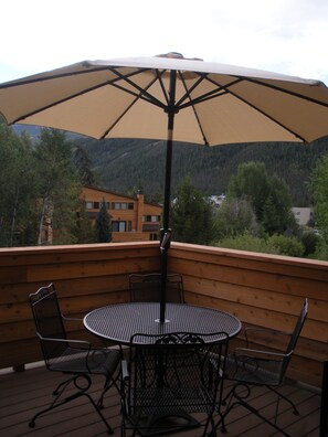 Eine Holzdielen-Terrasse mit einem runden Metalltisch und Stühlen, einem Sonnenschirm und Blick auf einen Berg sowie Gebäude im Hintergrund.