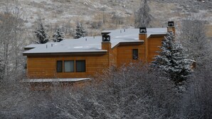 Eine Holzhütte mit schneebedecktem Dach, umgeben von Bäumen und einer hügeligen Landschaft.
