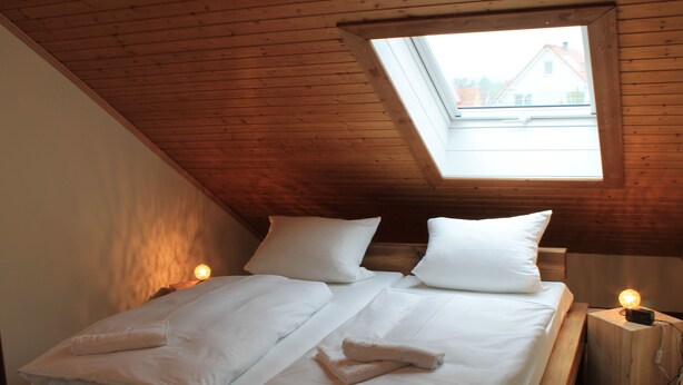 Ein Schlafzimmer mit schrägem Dach, einem Oberlicht, einem Bett mit weißen Leinen, zwei Nachttischlampen und einem Holznachttisch.