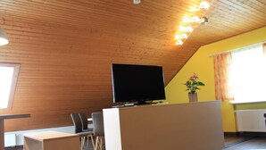 Ein Zimmer mit Holzboden, einem Flachbildfernseher auf einem weißen Schrank, einer Topfpflanze und einem Fenster mit Vorhängen.