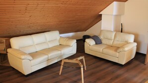 Ein Zimmer mit holzverkleideter Schräge, einem weißen Ledersofa, einem kleineren Sessel und einem Glastisch.