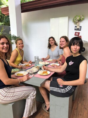 Un groupe de personnes est assis autour d’une table, devant des assiettes de nourriture. On aperçoit un ventilateur et, sur le mur, un panneau sur lequel est écrit le mot « MAISON ».