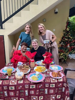Un groupe de personnes est assis à une table à l’extérieur, devant une variété de plats et de boissons. On aperçoit un sapin de Noël en arrière-plan, et un escalier mène à un étage supérieur.