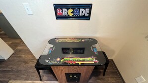 Ein Retro-Arcade-Automat mit Holzfuß und farbenfroher Grafik, platziert in einem Raum mit Holzboden und einem an der Wand befestigten Schild darüber.
