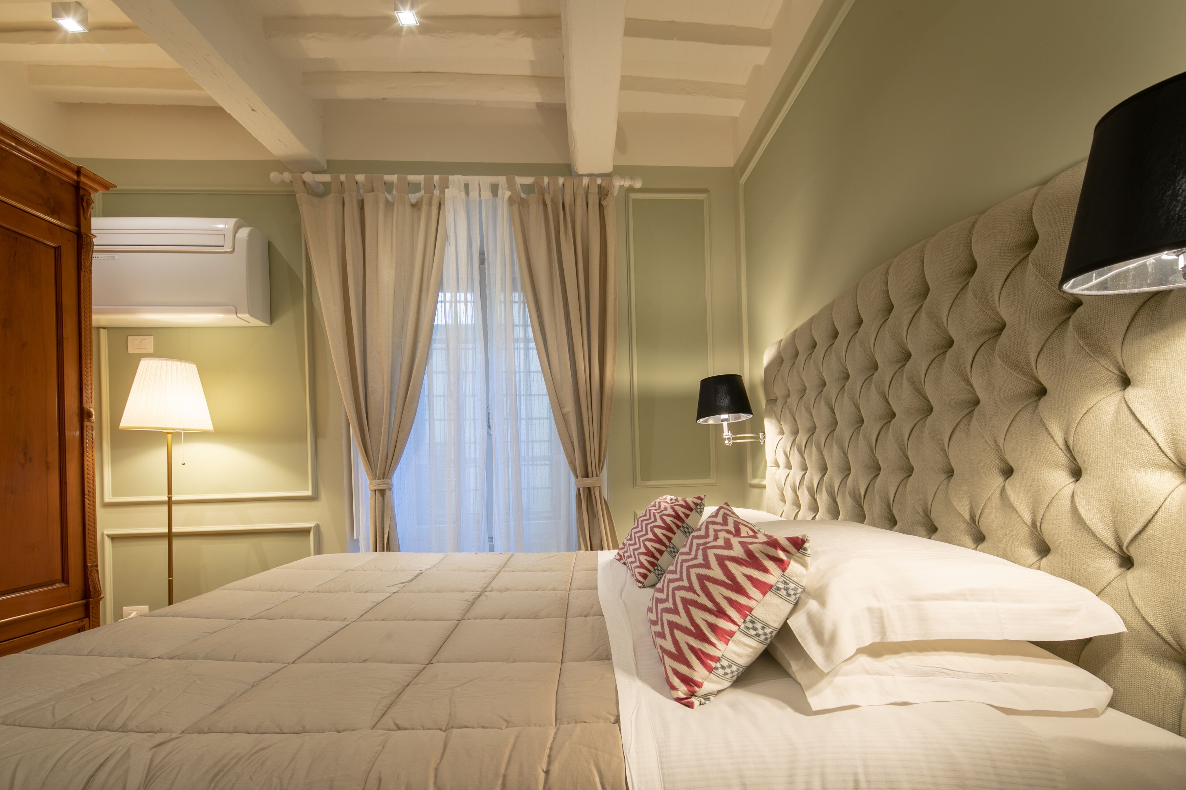 Habitación de hotel con una cama grande, cabecero, lámpara, equipo de aire acondicionado y ventana con cortinas.