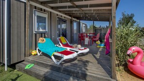 Een overdekt buitengebied met houten terras, twee ligbedden, een roze flamingo drijvend object en een patio tafel met stoelen.