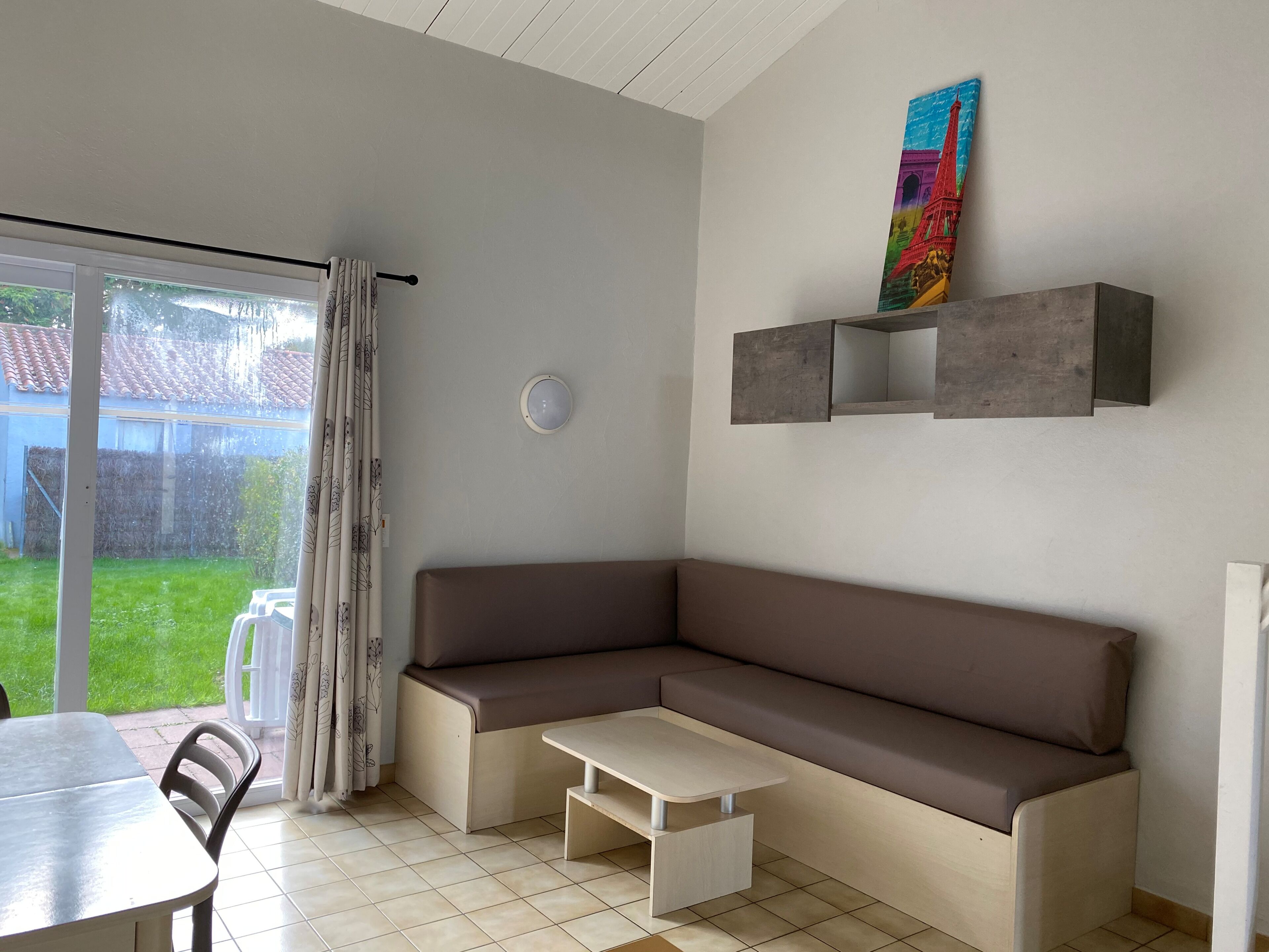 Un salon moderne avec un canapé d’angle marron, une petite table blanche et un coin repas composé d’une table et de chaises. Un tableau coloré représentant une tour est accroché au mur, et une porte-fenêtre coulissante donne accès à un jardin.