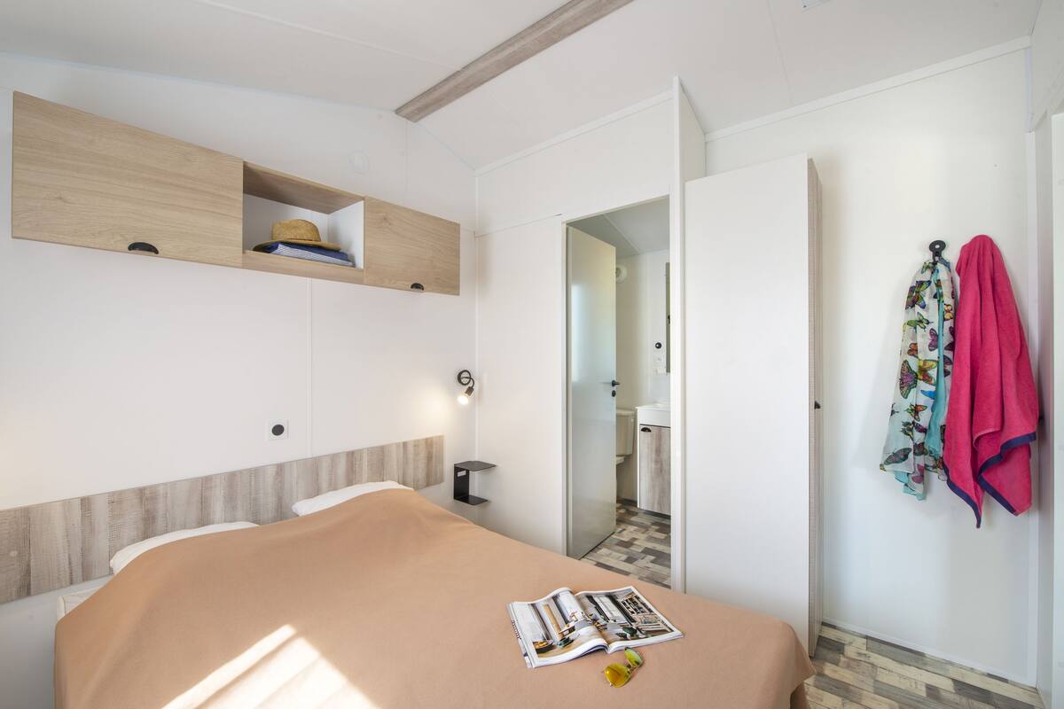 Une chambre à coucher comprenant un lit, une étagère en bois, un miroir et une porte donnant sur une salle de bain.