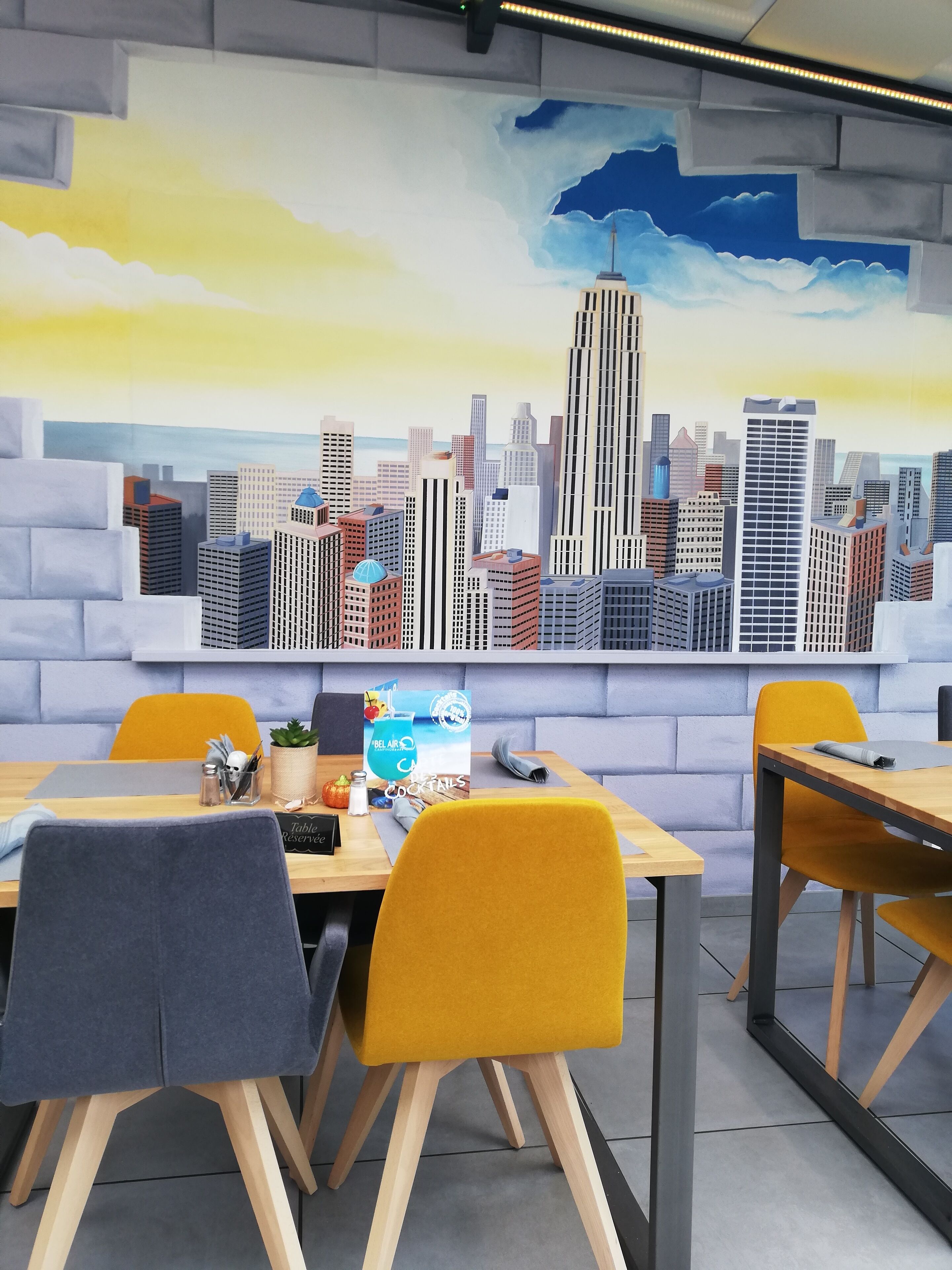 A modern dining area with a cityscape mural, wooden tables, and yellow chairs.