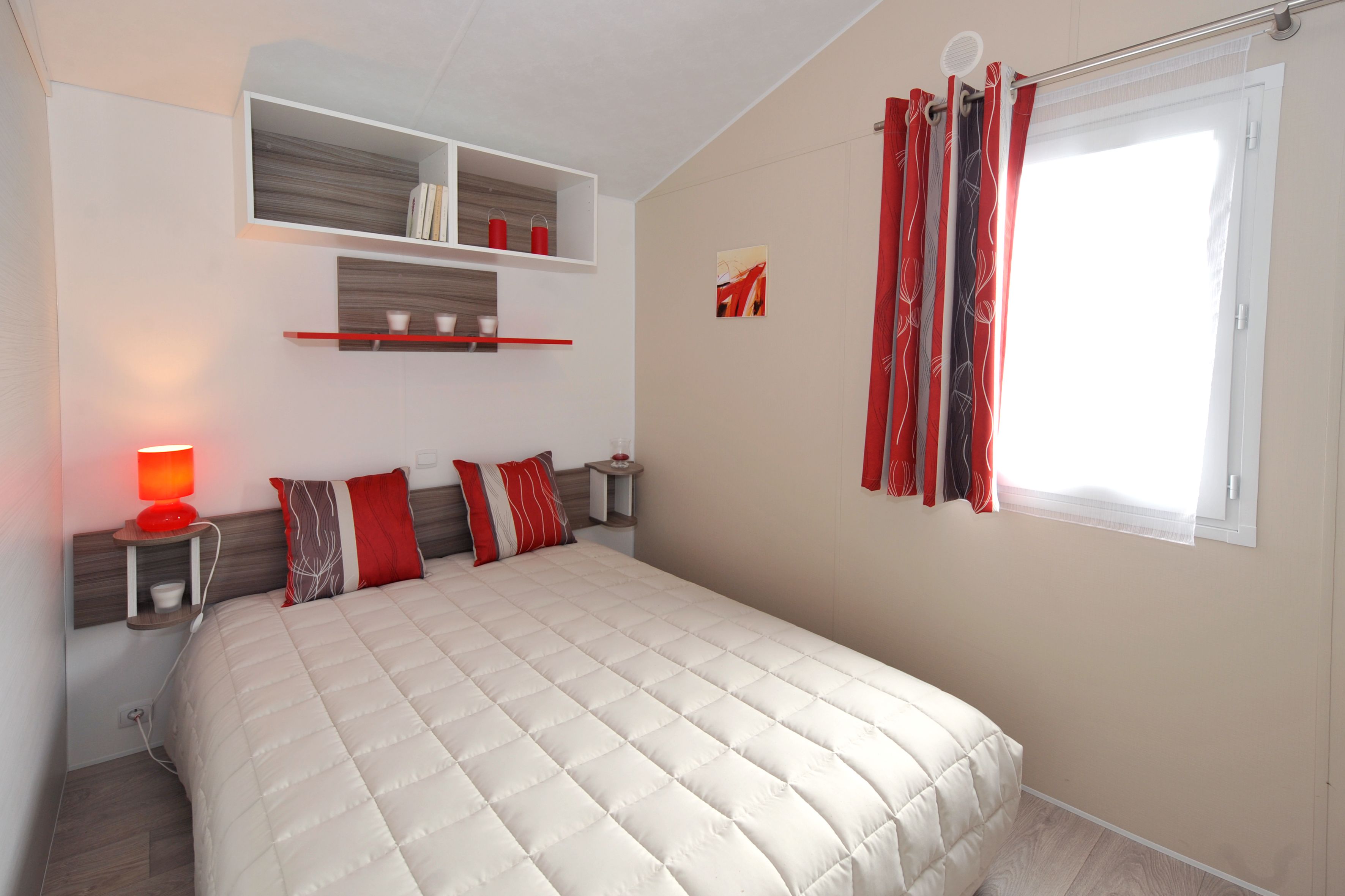 A bedroom with a large bed, a bedside table with a lamp, a shelf above the bed, and a window with curtains.