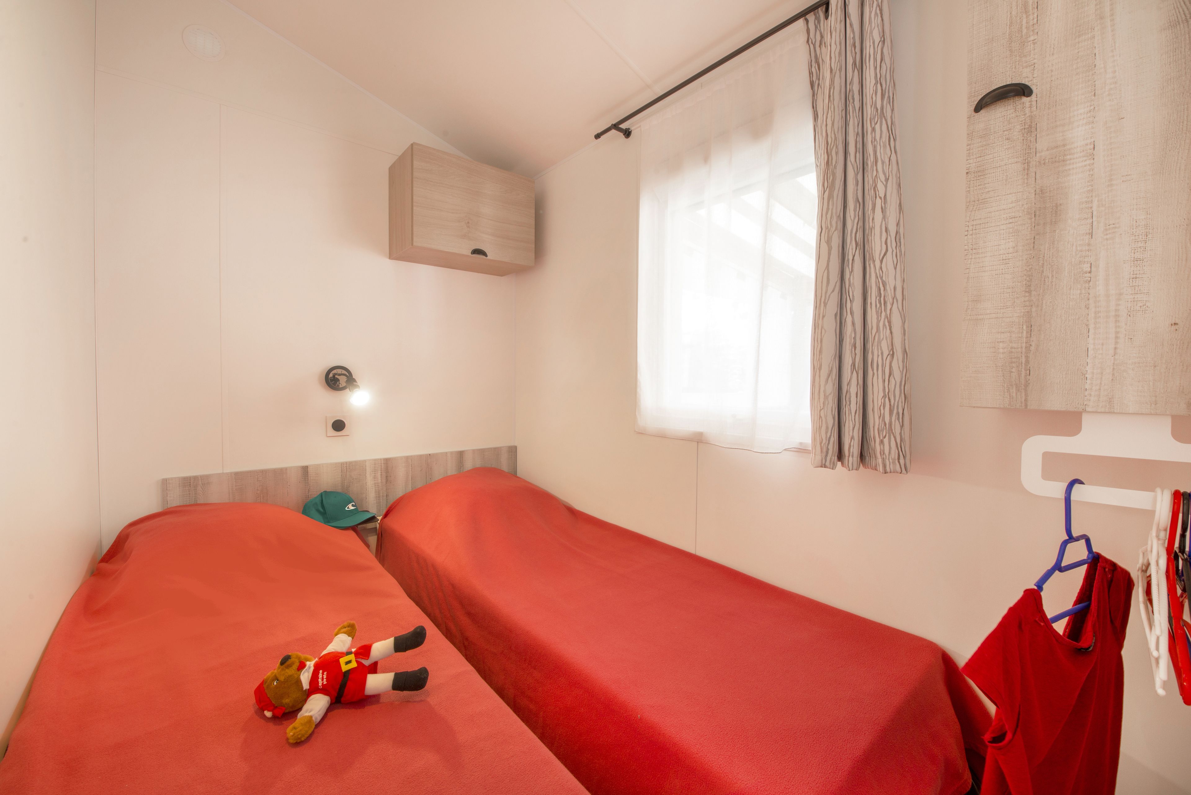 Una cama individual con colcha roja, un pequeño armario de madera, una lámpara de pared y una ventana con cortinas.