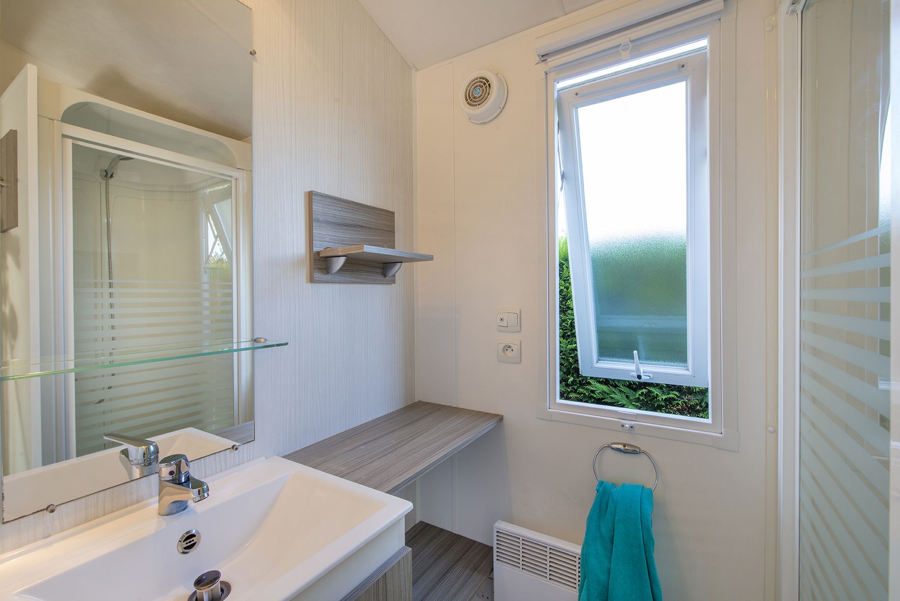 Een badkamer met een glazen douchecabine, een witte wastafel en een raam met uitzicht op groen.
