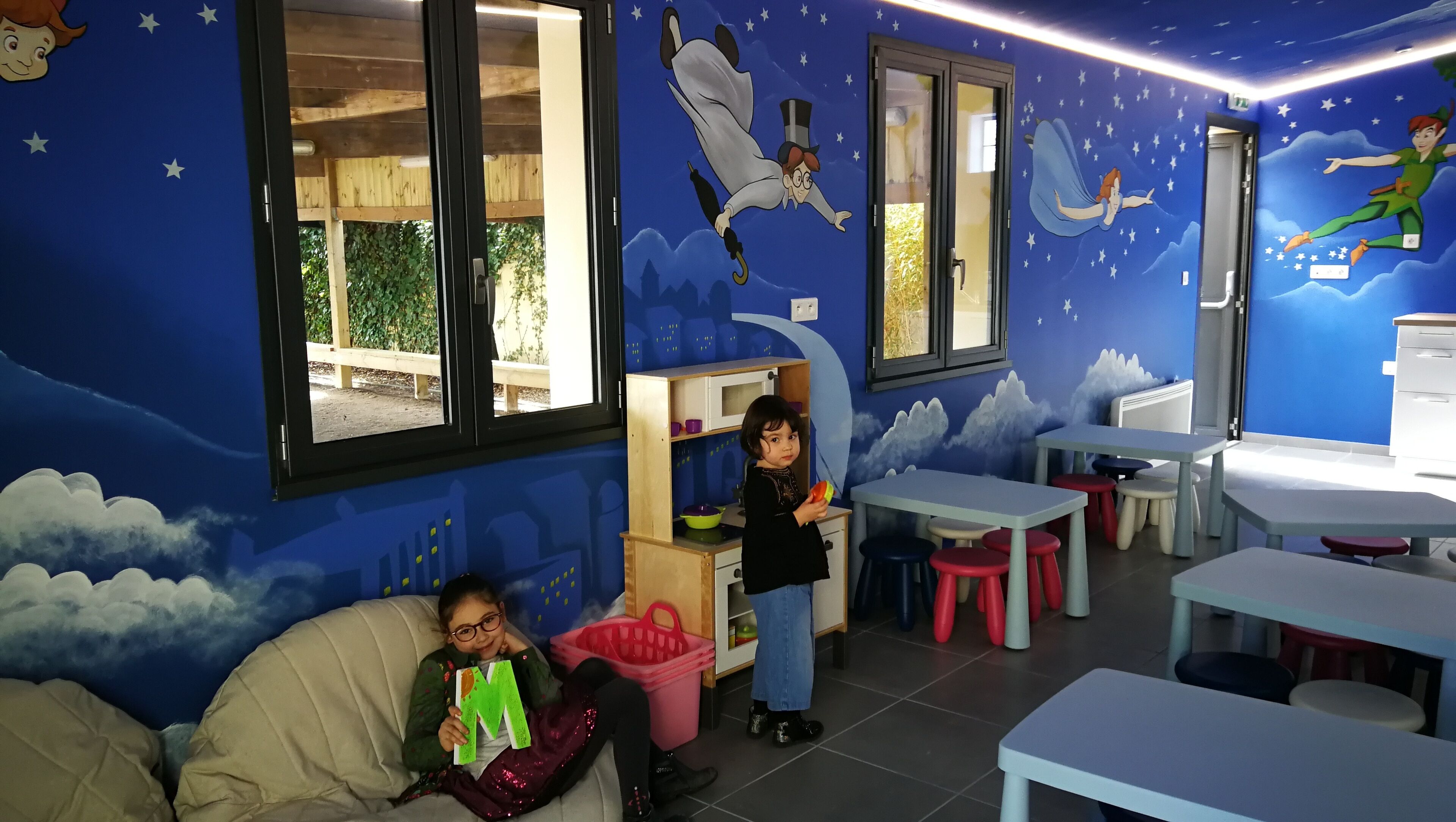 Une salle de jeux pour enfants décorée dans des tons bleus, avec une grande fenêtre, un espace de jeu équipé de tables et de chaises, et une fresque murale représentant un personnage volant.