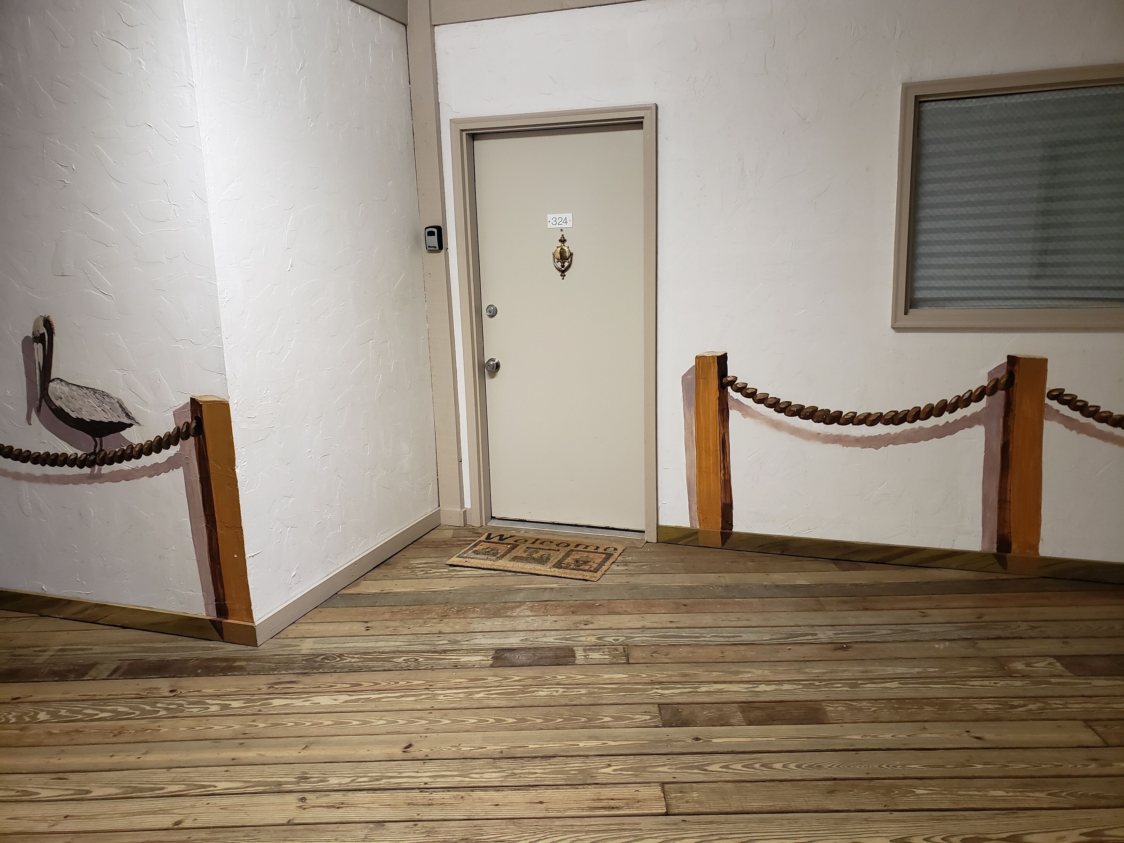 Um corredor com piso de madeira, uma porta branca com placa de identificação e um poste de madeira com uma barreira de corrente.