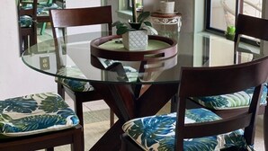 Un coin repas avec une table en verre entourée de chaises en bois dotées de coussins à motifs, un vase avec des fleurs blanches et un miroir accroché au mur.