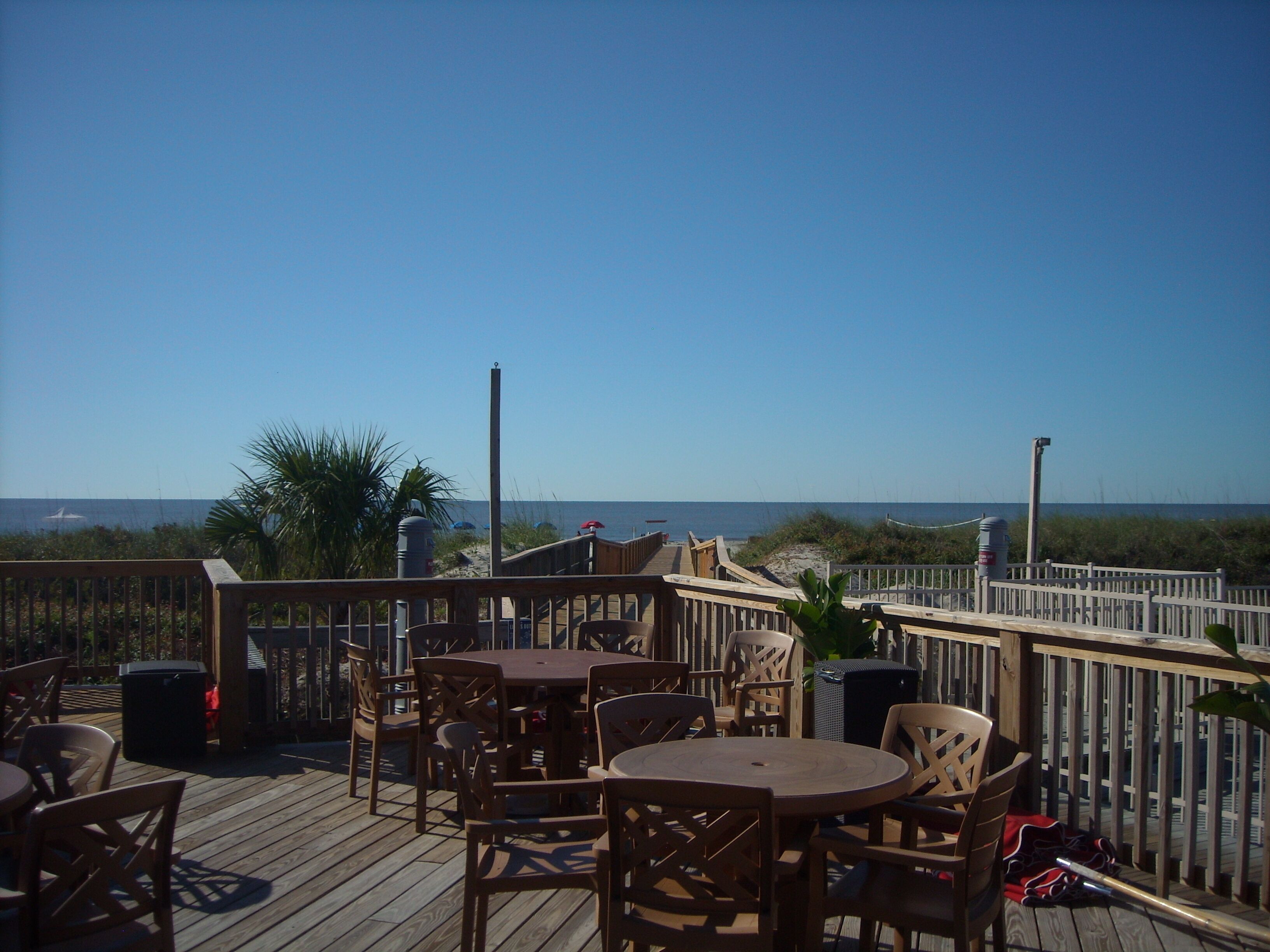 Een terras buiten met houten tafels en stoelen, met uitzicht op een strand en de oceaan op de achtergrond.