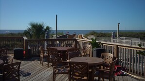 Une terrasse extérieure avec des tables et des chaises en bois, donnant sur une plage et offrant une vue sur l’océan en arrière-plan.