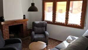 En stue med en murstenspejs, en læderlænestol, en sofa og et vindue med blondegardiner.