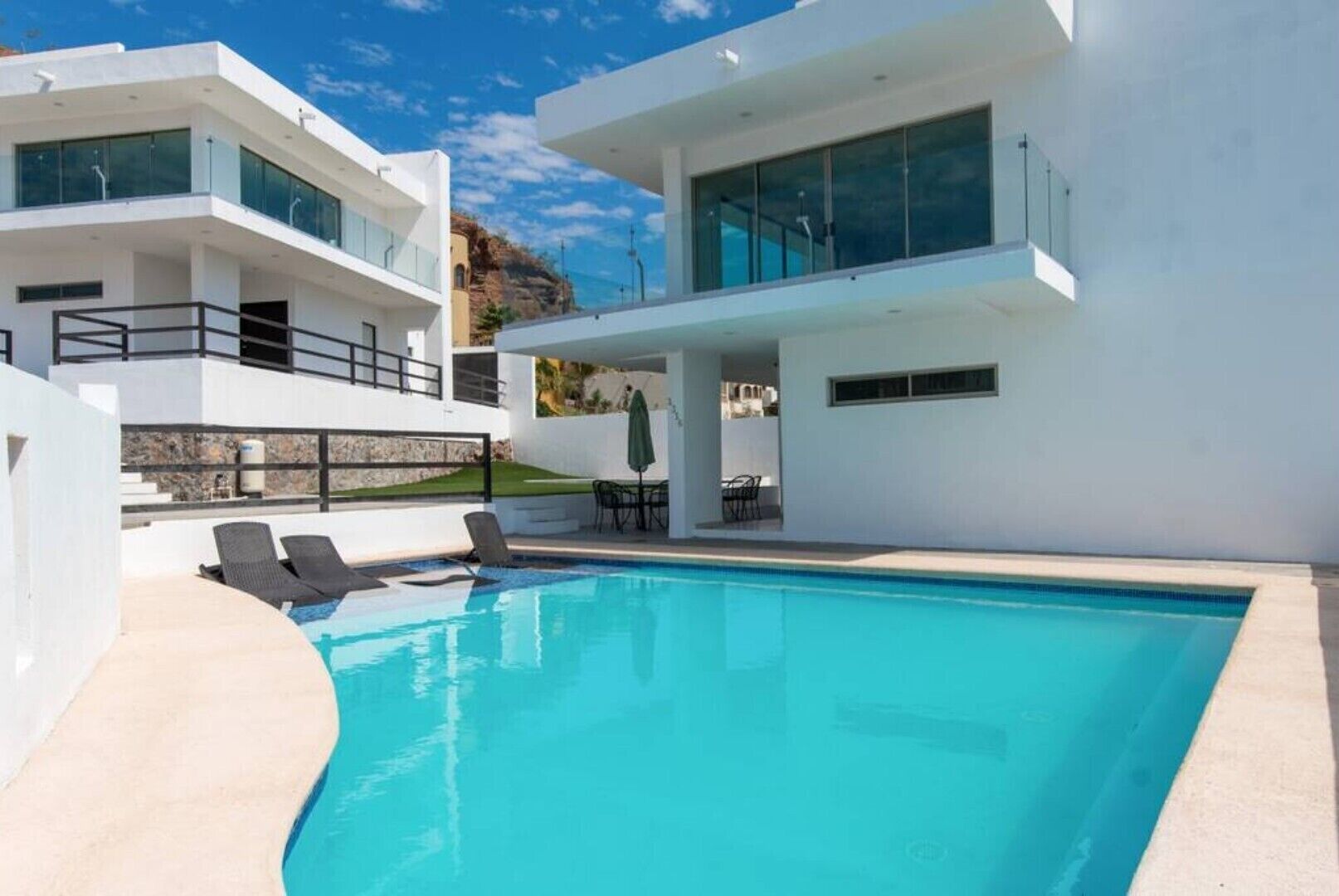 A modern two-story house with a swimming pool, outdoor furniture, and a view of the surrounding landscape.