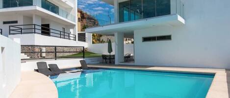 A modern two-story house with a swimming pool, outdoor furniture, and a view of the surrounding landscape.