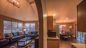 Amplia sala de estar con un sofá de cuero oscuro, una mesa de centro y un televisor sobre un mueble de madera. Hay una zona de comedor con una mesa y sillas, y ventanas amplias que proporcionan luz natural.