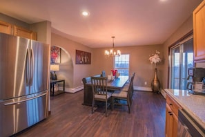 A modern kitchen with a stainless steel refrigerator, wooden dining table, and chairs. There is a chandelier above the table and a window with a view. - Hilltop Lodge (Beach Lake)