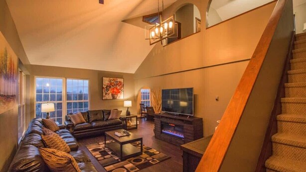 Amplia sala de estar con un sofá grande, una mesa de centro y una chimenea. Hay un televisor y una escalera que conduce a un nivel superior.