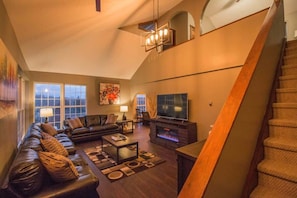 A spacious living room with a large sofa, a coffee table, and a fireplace. There is a television and a staircase leading to an upper level. - Hilltop Lodge (Beach Lake)