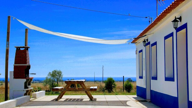 Un patio con mesa y sillas de madera, un horno de ladrillo y vista al mar.