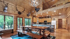 Interior de uma cabana de madeira com uma grande mesa de jantar, bancos e uma área de cozinha.