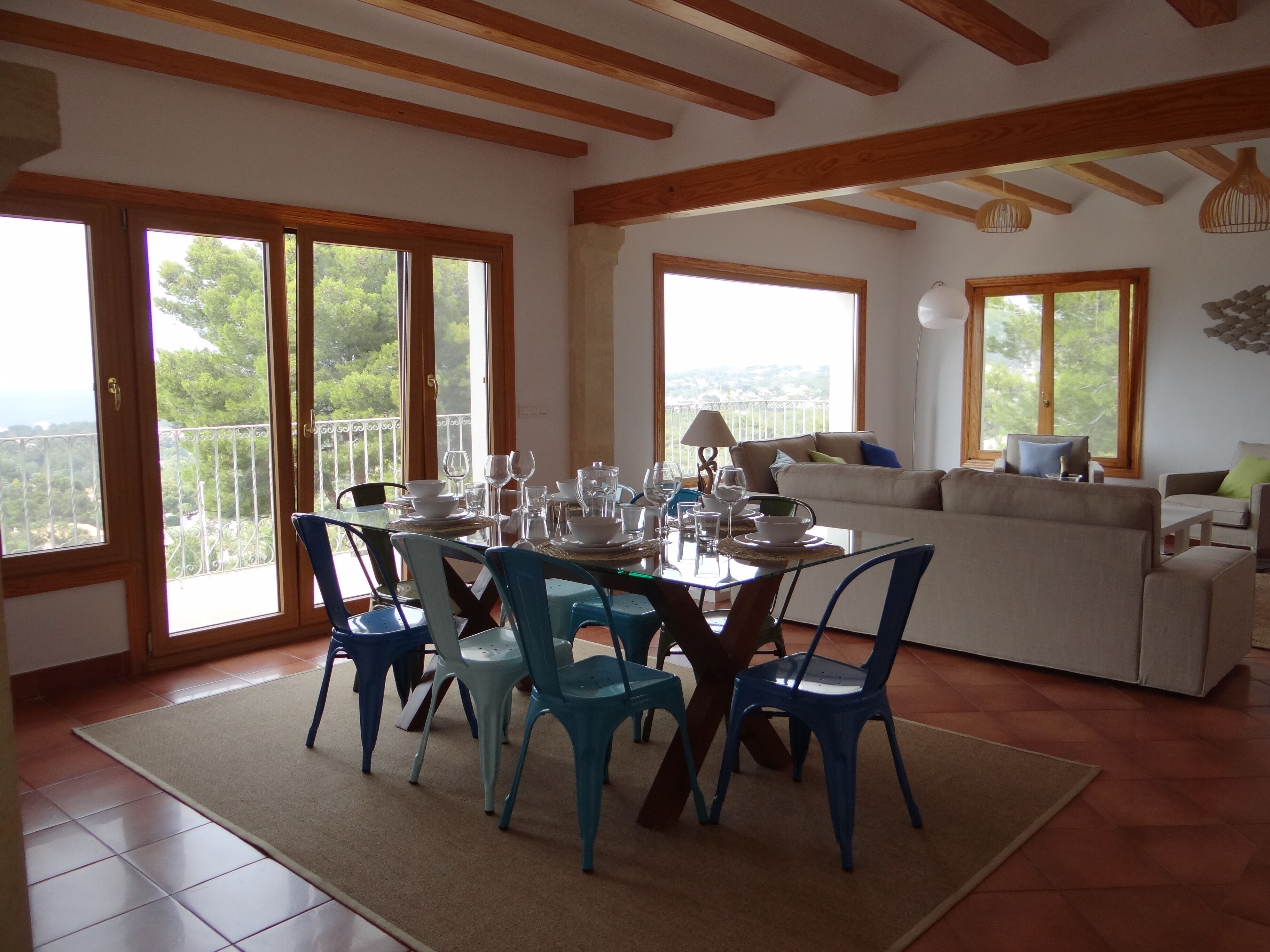 A dining area with a table set for six, surrounded by blue chairs, a sofa, and a balcony with a view.