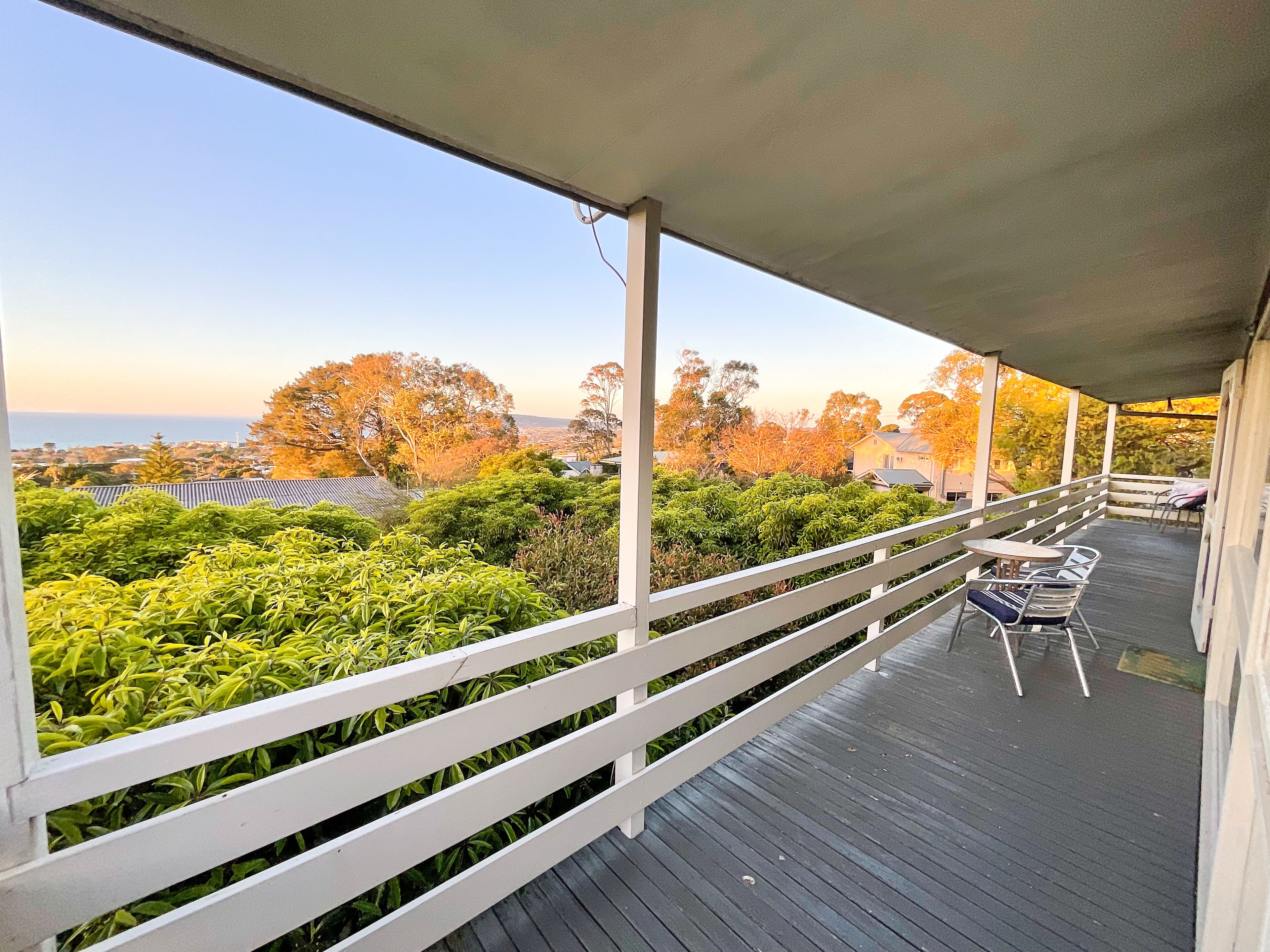 A covered outdoor area with a wooden deck, white railing, and a view of the sea and trees.