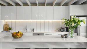 A modern kitchen with white cabinetry, a central island with a fruit bowl, and a vase with flowers. - Pet Friendly - Danny Boy Coastal Retreat in Rye (Rye)