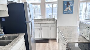 Een moderne keuken met witte kasten, een zwarte koelkast, een magnetron en een kookplaat. Op de vloer ligt een blauw vloerkleed en er is een raam met uitzicht.