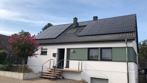 A modern house with solar panels on the roof, a staircase leading to the entrance, and a small garden with pink flowers. - Bed 'n Riesling - The vacation apartment in Forst a.d. Wine route with its own garden (Forst an der Weinstraße)